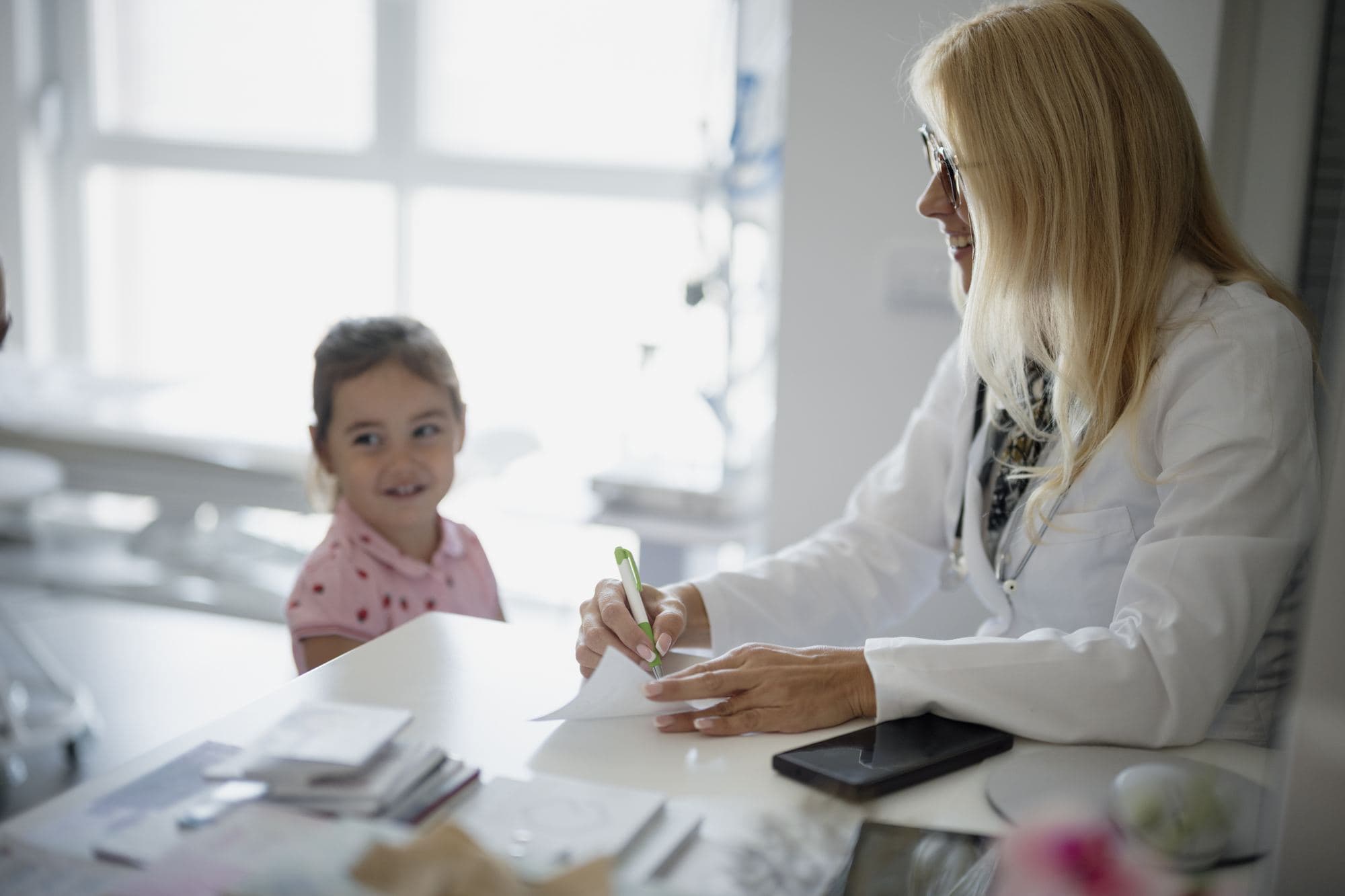 A doctor writes at her desk with a young girl sitting opposite