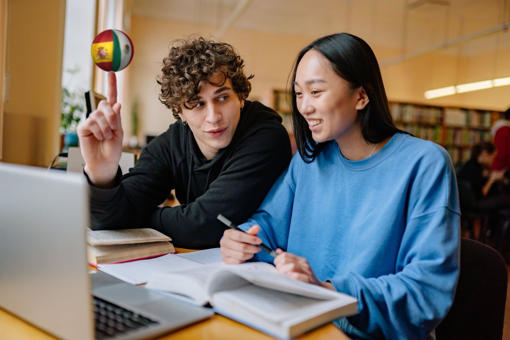 Two students go over a book and a laptop together in a university library; he balances a ball with Mexico's flag merged with Spain's on the tip of his index finger.