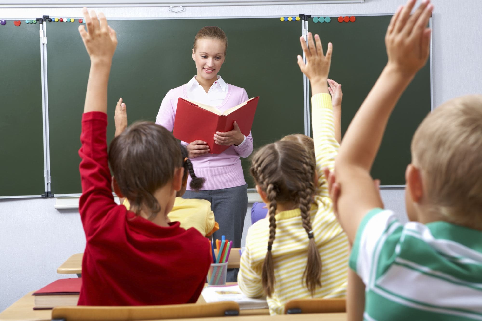Children raise their hands in class while the teacher reads by the blackboard