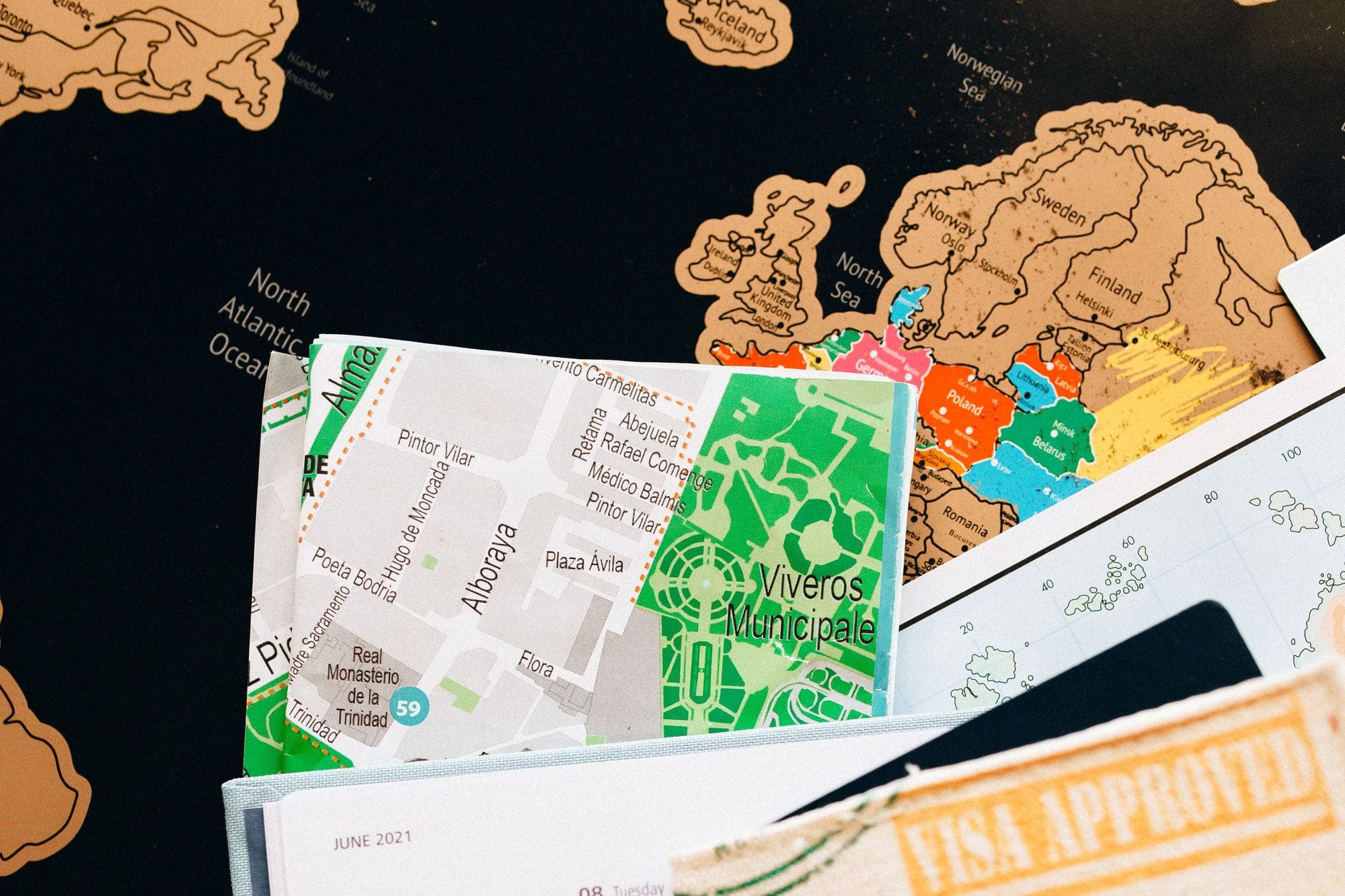 A street map of Madrid and a scratch-off map of Europe on a dark table, next to a passport stamped «Visa approved»