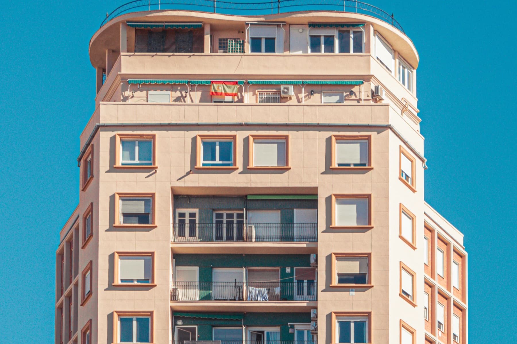 A pink-toned apartment building with balconies and a Spanish flag, against a deep blue sky
