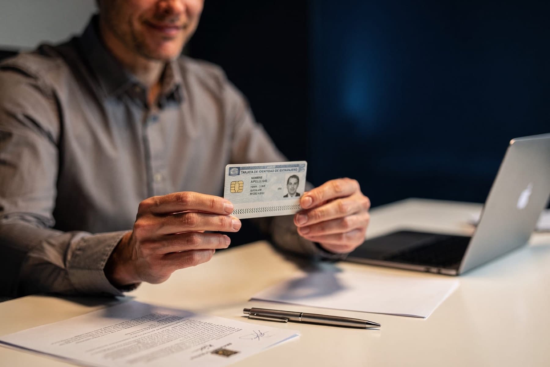 A man holds up a residence card in front of a laptop and some papers on the table