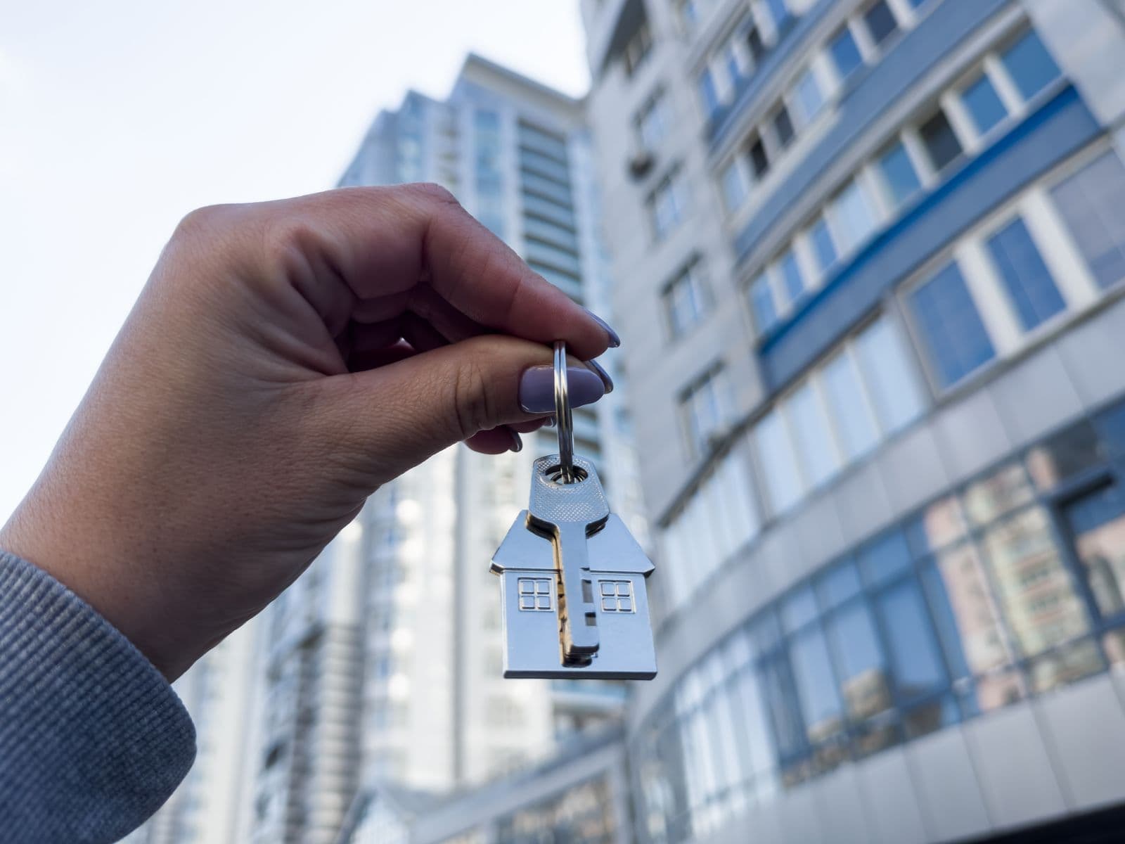 A hand holding keys on a house-shaped keyring in front of an apartment building